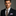 Man wearing a black suit with a patterned tie and purple pocket square on a gray background