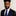 Man wearing a black suit with a blue tie and pocket square against a neutral background