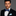 Man wearing a black suit with a blue plaid bow tie and light blue handkerchief on a gray background