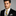 Man wearing a black suit with a yellow tie against a gray background