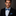 Man wearing a black suit with a blue checkered bow tie and pocket square against a dark background