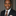 Man wearing a dark suit with a brown tie and pocket square against a neutral background