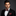Man wearing a black suit with a red and black plaid bow tie and pocket square on a dark background