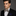 Man wearing a black suit with a patterned brown floral and stripe bow tie and accenting blue and brown pocket square on a gray background
