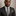Man wearing a dark suit with a patterned green and burgundy paisley tie and pocket square against a neutral background