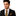 Man wearing a black suit with a yellow gold and black geometric small diamonds patterned tie and pocket square on a white background