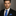 Man wearing a black suit with a blue and grey check tie and pocket square against a gray background