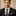 Man wearing a black suit with a blue and brown striped tie and pocket square against a beige background