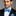Man wearing a black suit with a light blue bow tie and matching pocket square on a gray background