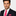 Man wearing a dark suit with a white shirt and pink tie and pocket square on a light gray background