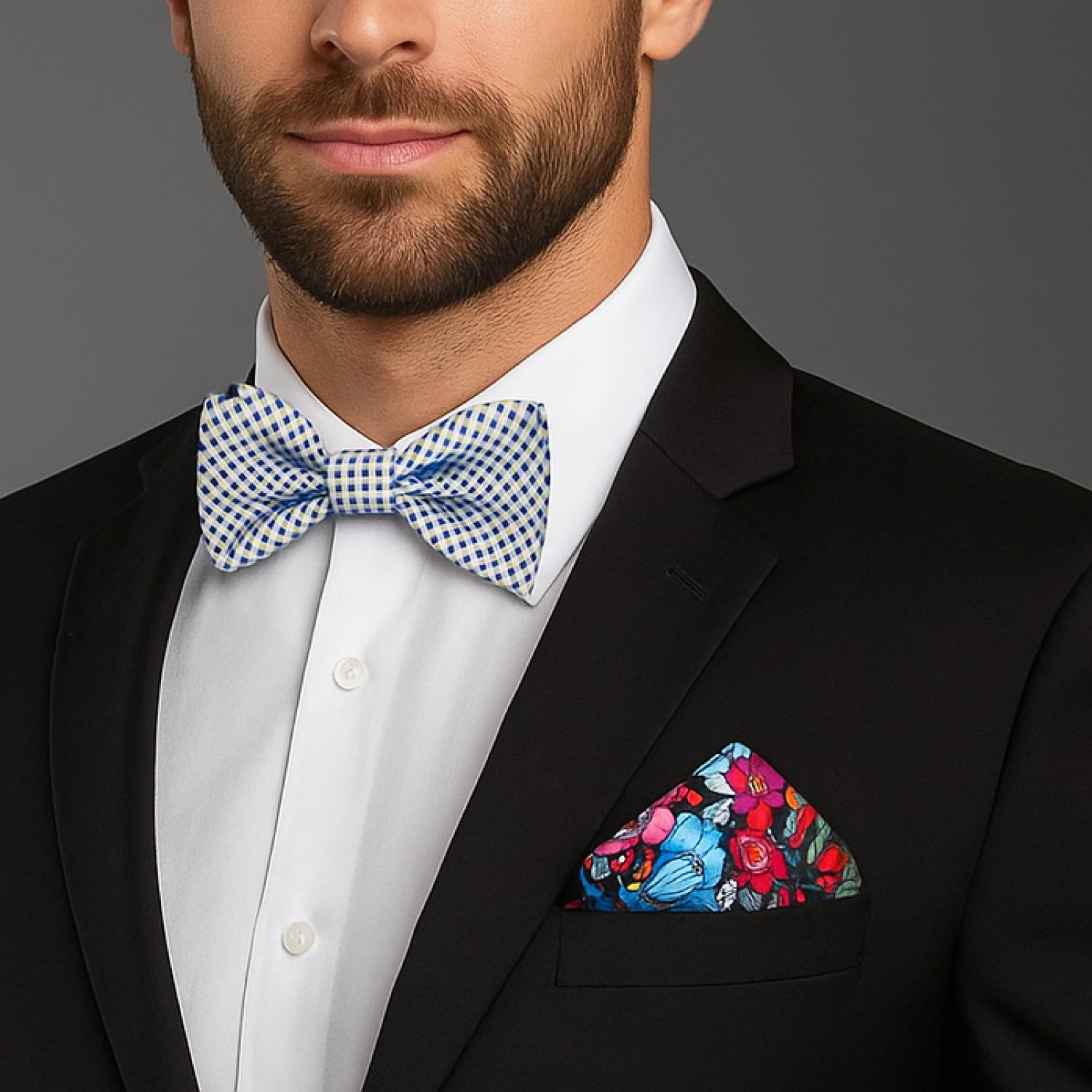 Man wearing a black suit with a white shirt, blue checkered bow tie, and colorful floral pocket square on a gray background.