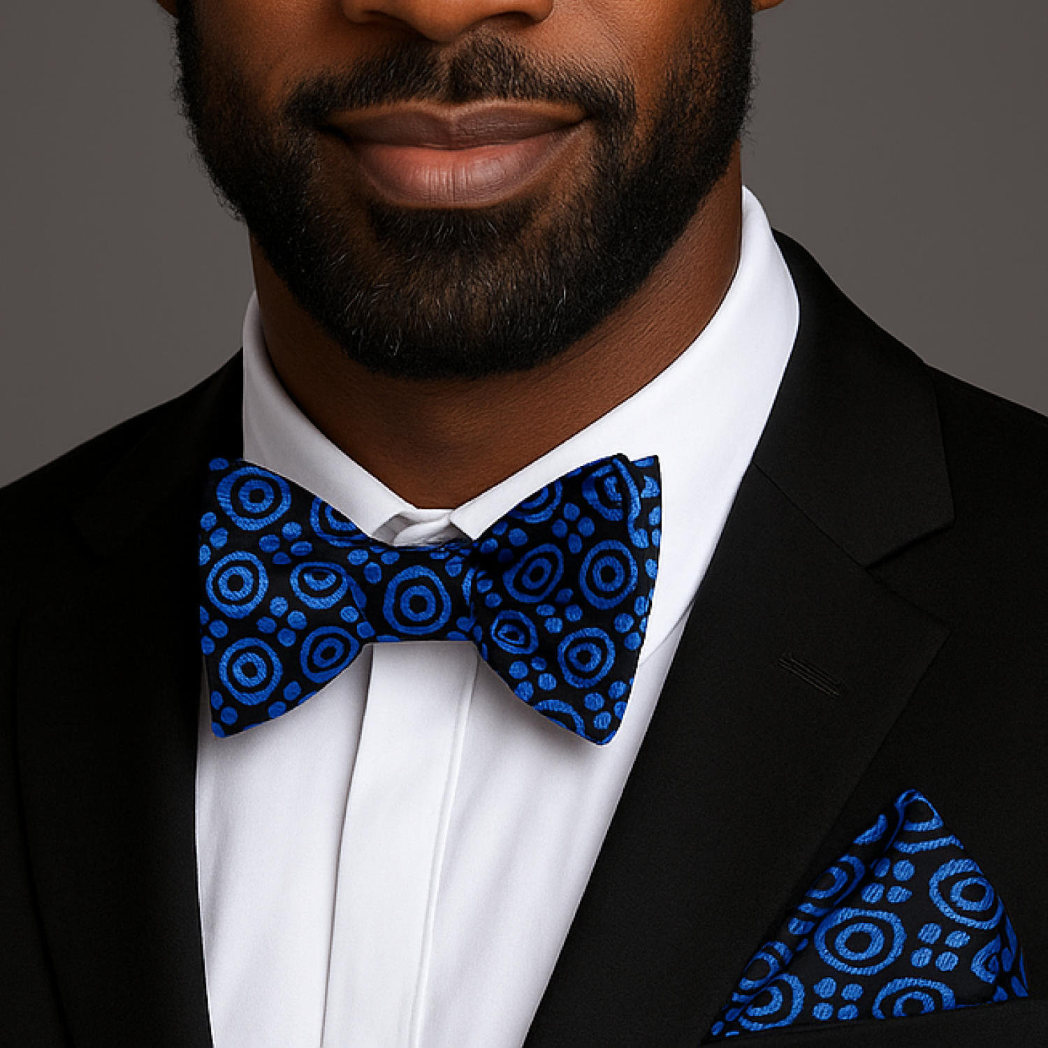 Man wearing a black suit with a blue patterned bow tie and pocket square against a gray background