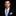 Man wearing a black suit with a blue striped thin tie and pocket square on a dark background