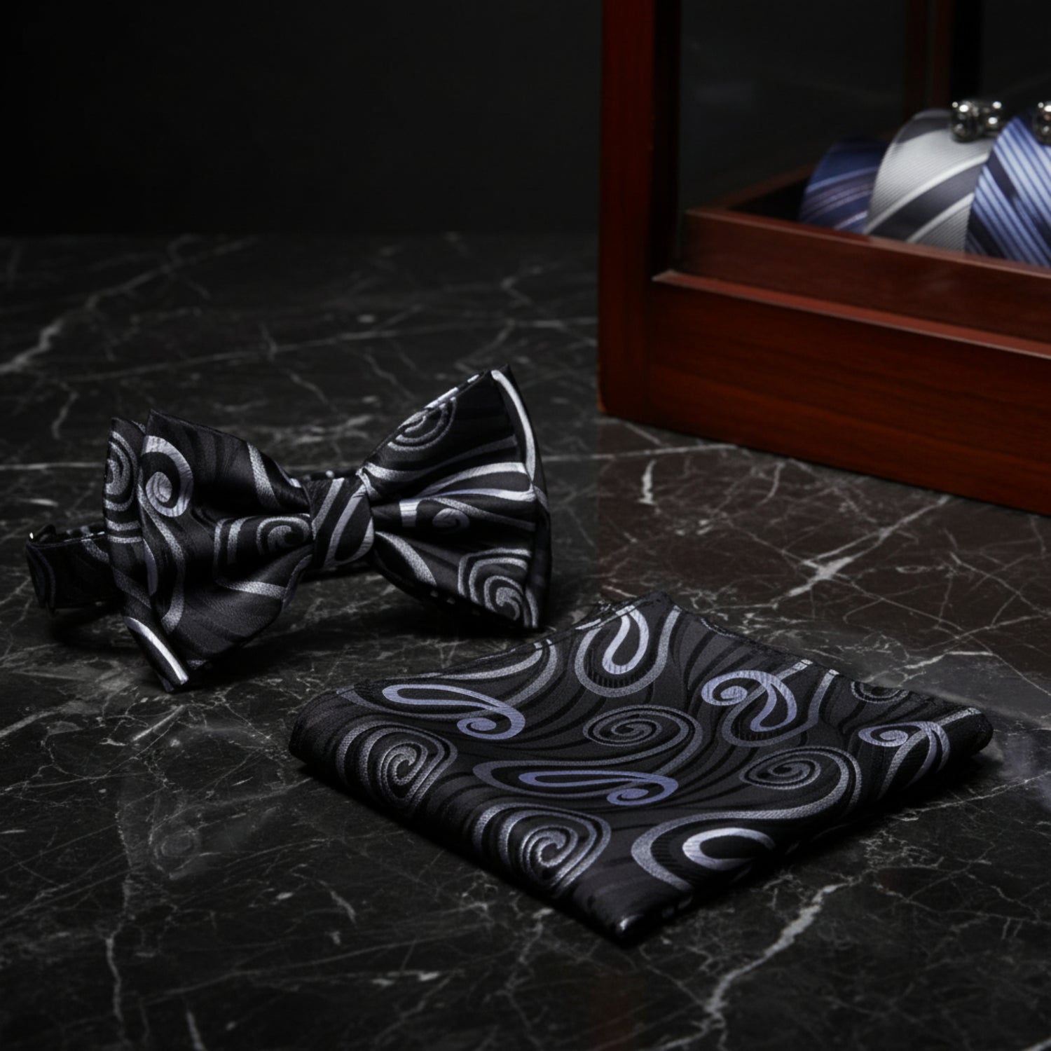 A matching men's formal accessory set featuring a pre-tied bow tie and a folded pocket square, both in black with a swirling paisley pattern in silver and gray.