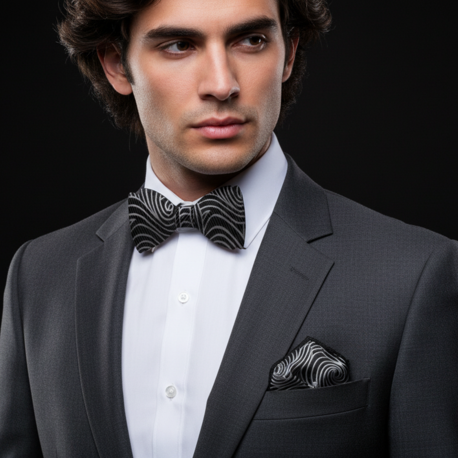 Man wearing a gray suit with a black and white patterned bow tie and pocket square on a black background