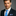 Man wearing a dark suit with a blue tie and pocket square on a gray background