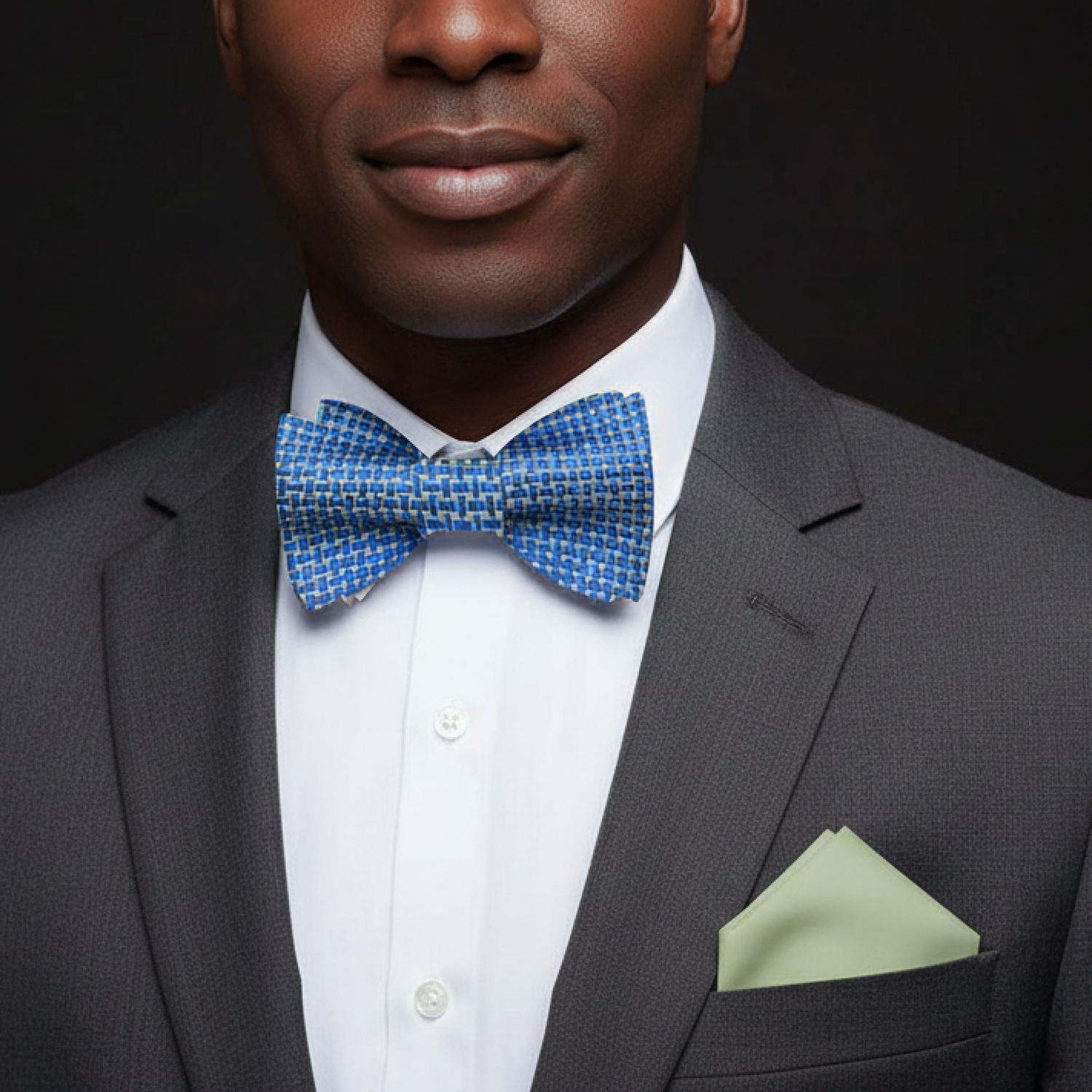 Man wearing a gray suit with a blue bow tie and green pocket square on a black background