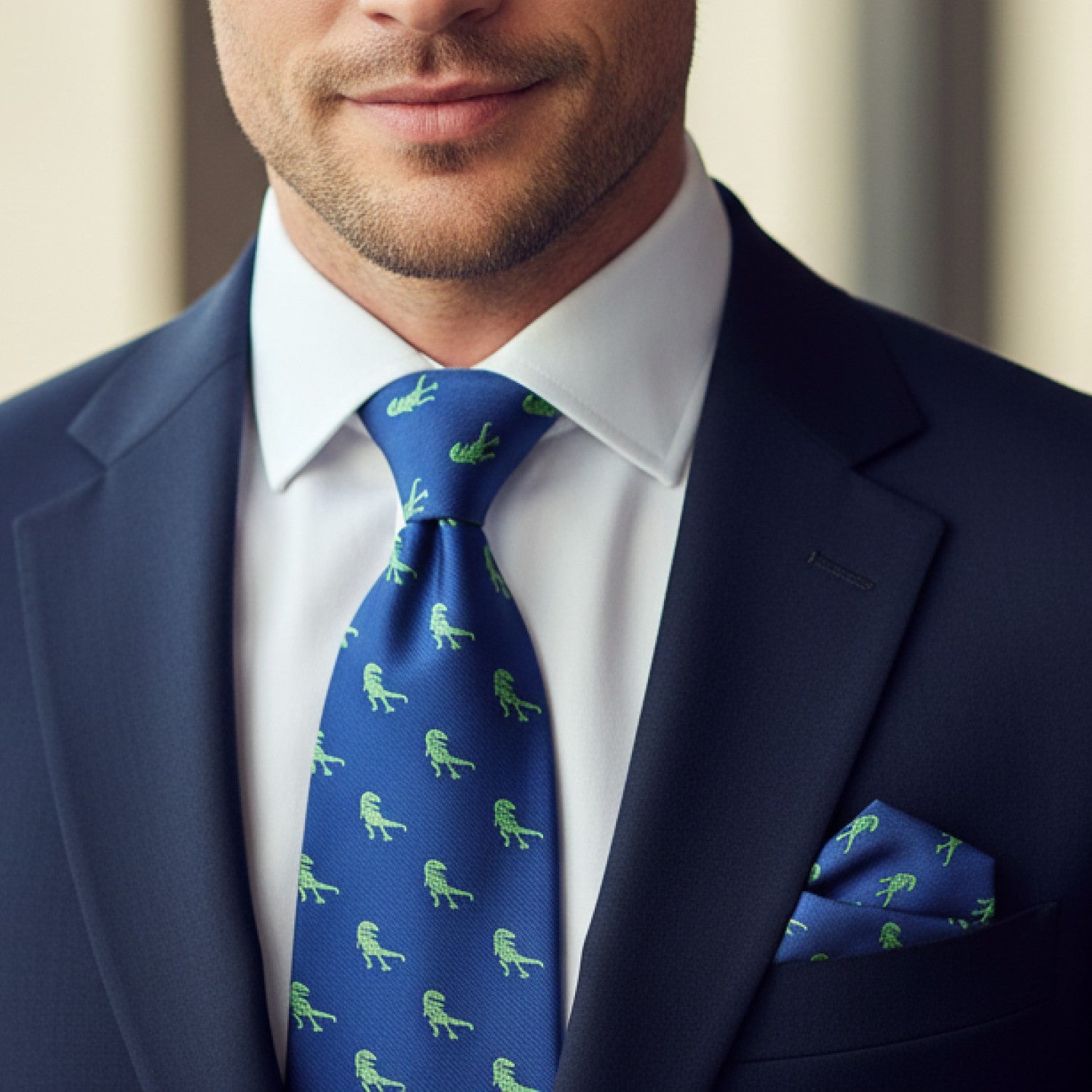 Man wearing a navy suit with a blue tie and matching pocket square featuring green dinosaur patterns.