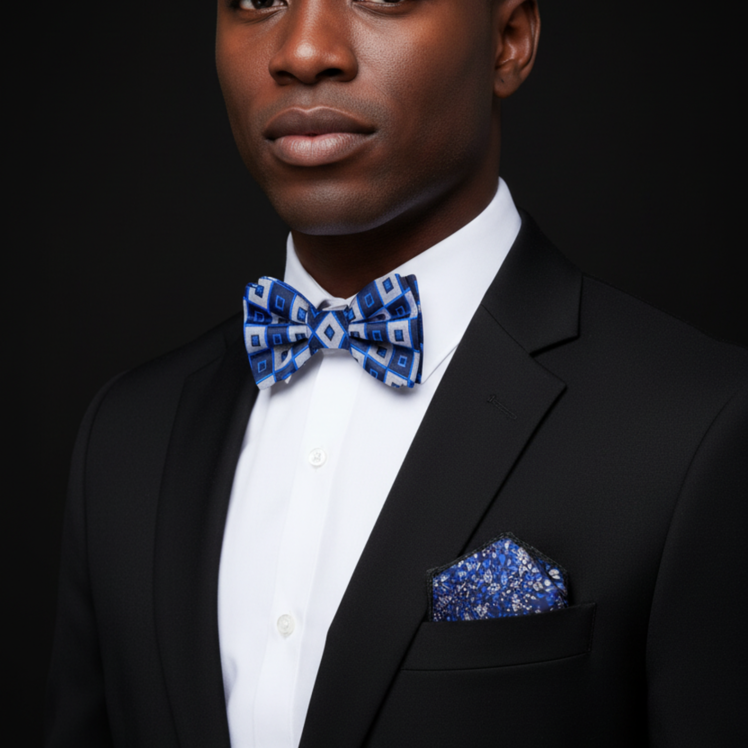 Man wearing a black suit with a white shirt and blue patterned bow tie and pocket square against a black background