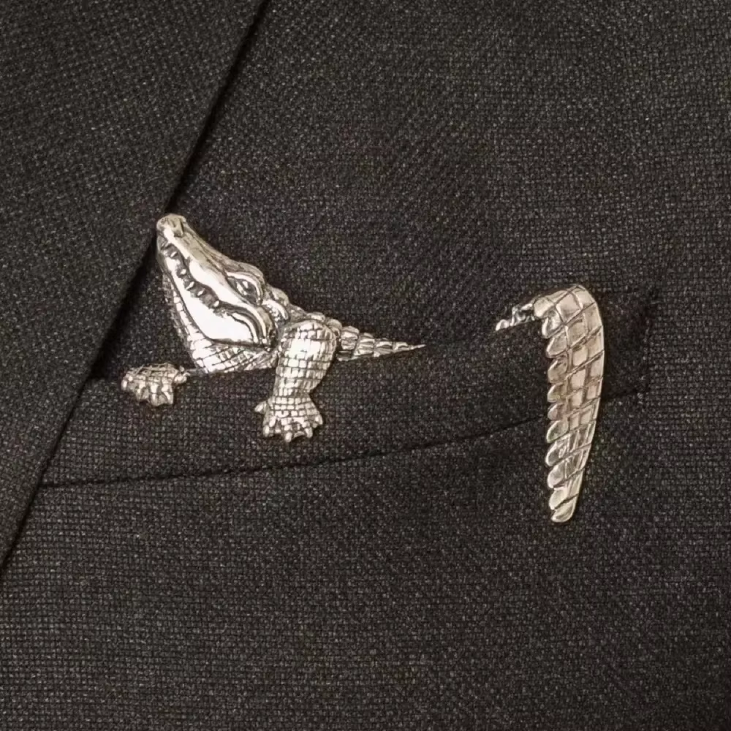A silver alligator-shaped pocket square or accessory pinned to a dark charcoal gray suit jacket lapel.