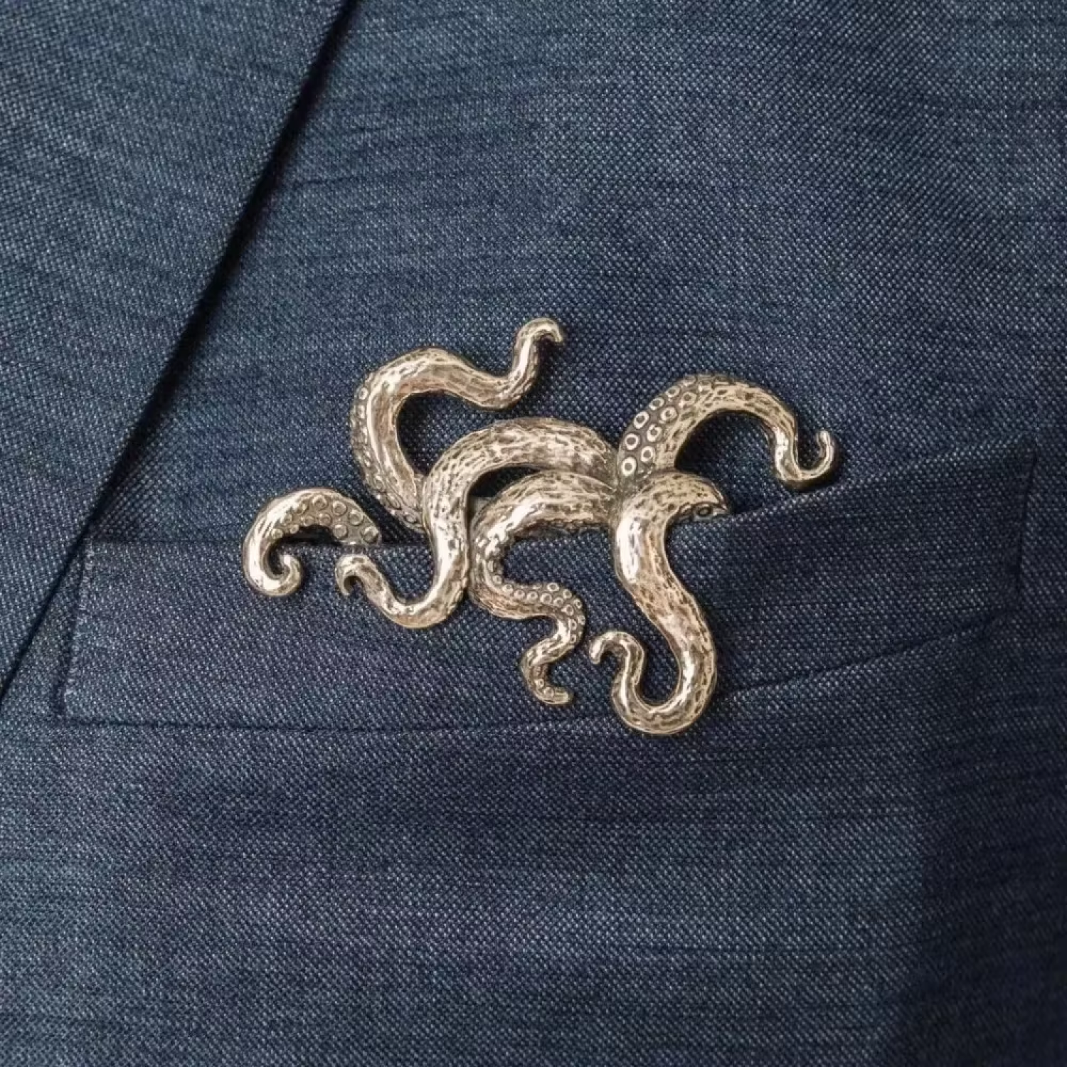 A silver-toned octopus tentacle brooch pinned to the chest pocket of a textured navy blue suit jacket.