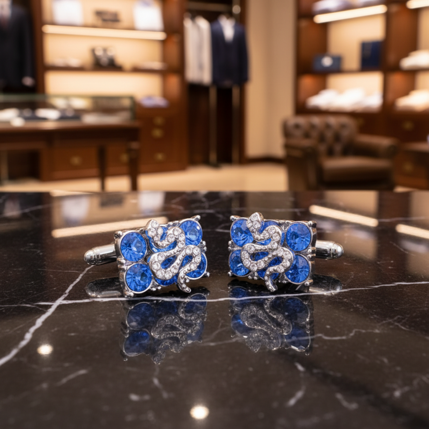 A pair of silver-toned square cufflinks decorated with faceted blue gemstones and clear rhinestone scrollwork, displayed on a reflective black surface.