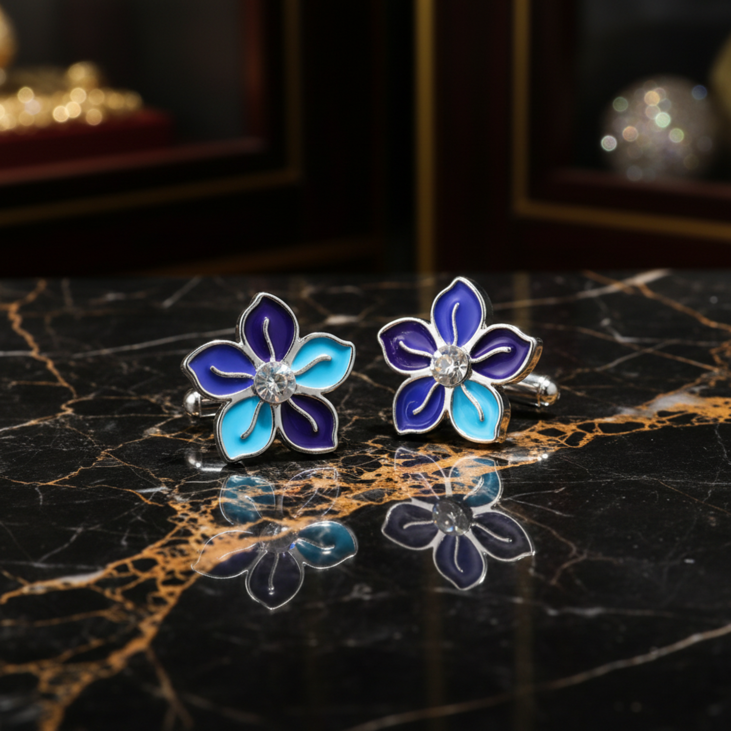 A pair of silver-tone flower stud earrings with vibrant blue and light blue enamel petals, set against a solid black background with a soft reflection below.