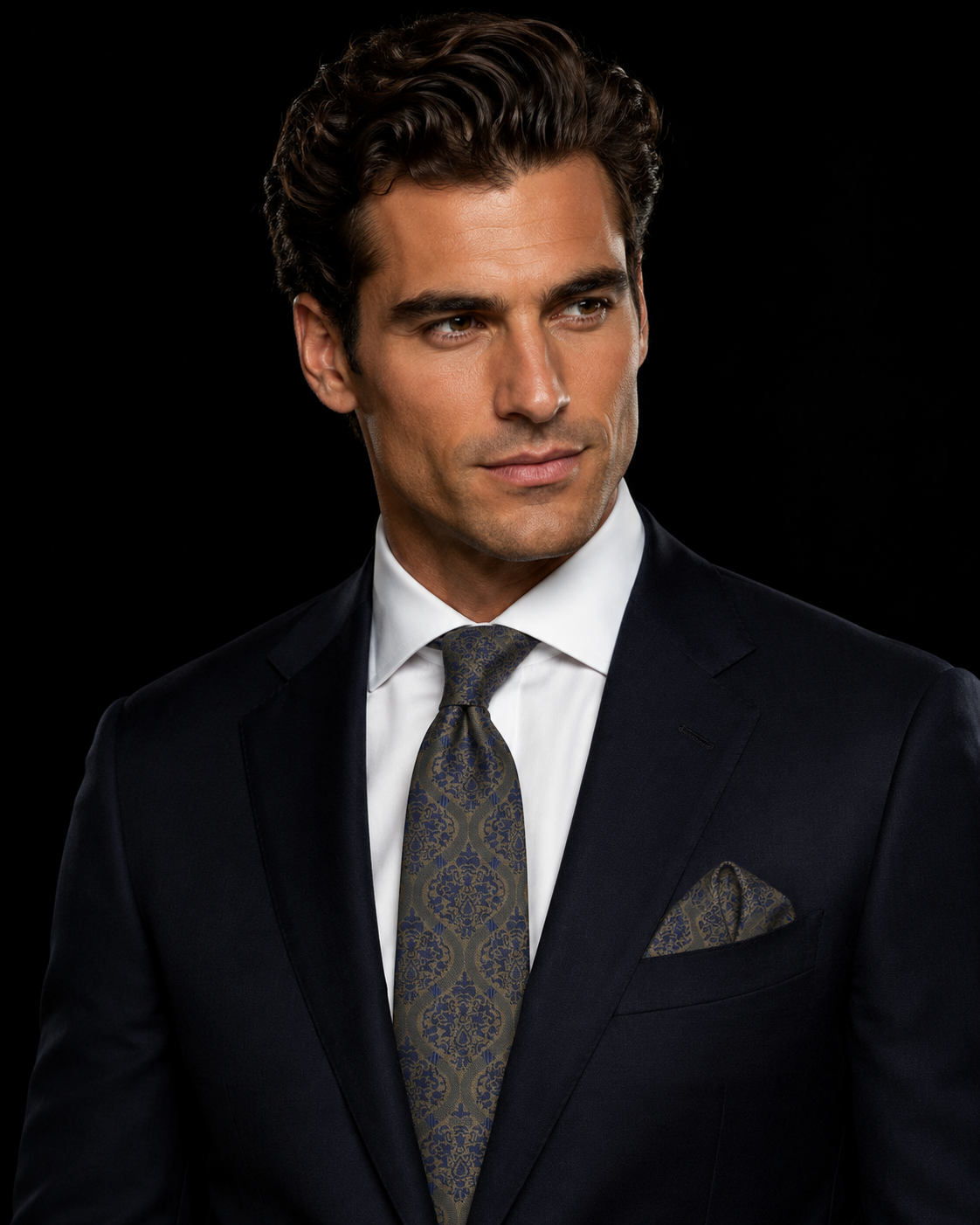 A man wearing a dark navy suit, white dress shirt, and a patterned blue and gold tie, set against a solid black background.