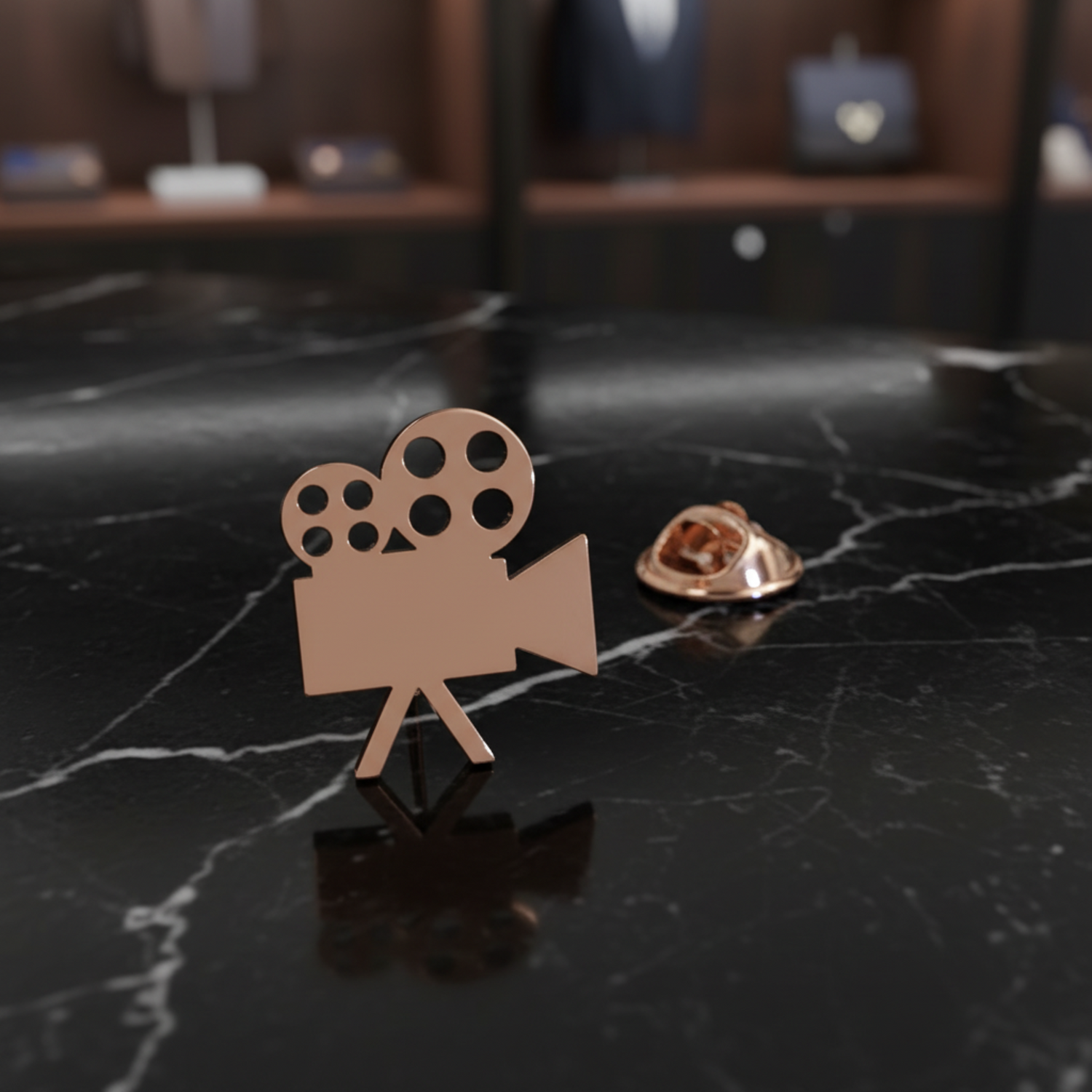 A rose gold metal pin shaped like a vintage film camera, shown alongside its circular metal backing on a textured black background.