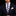 A man wearing a navy blue suit, white dress shirt, a blue and beige striped necktie, and a matching patterned pocket square against a black background.