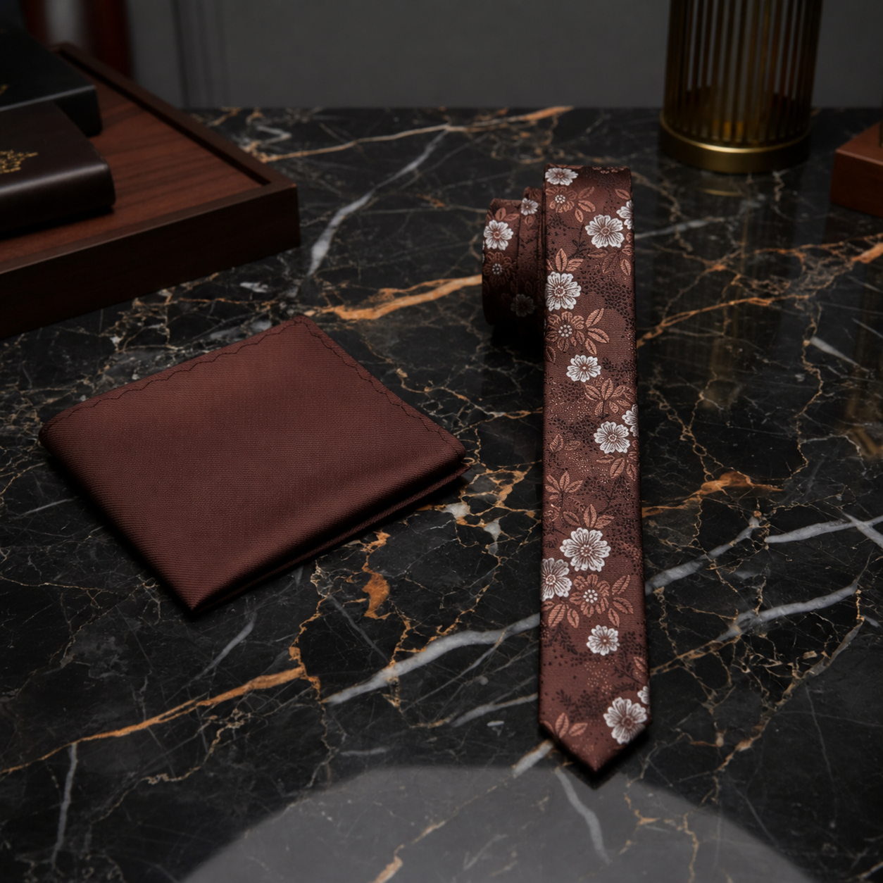 A men's formal accessory set featuring a brown necktie with a white floral pattern, a matching brown pocket square, and a rolled tie, arranged on a black and gold marble surface.