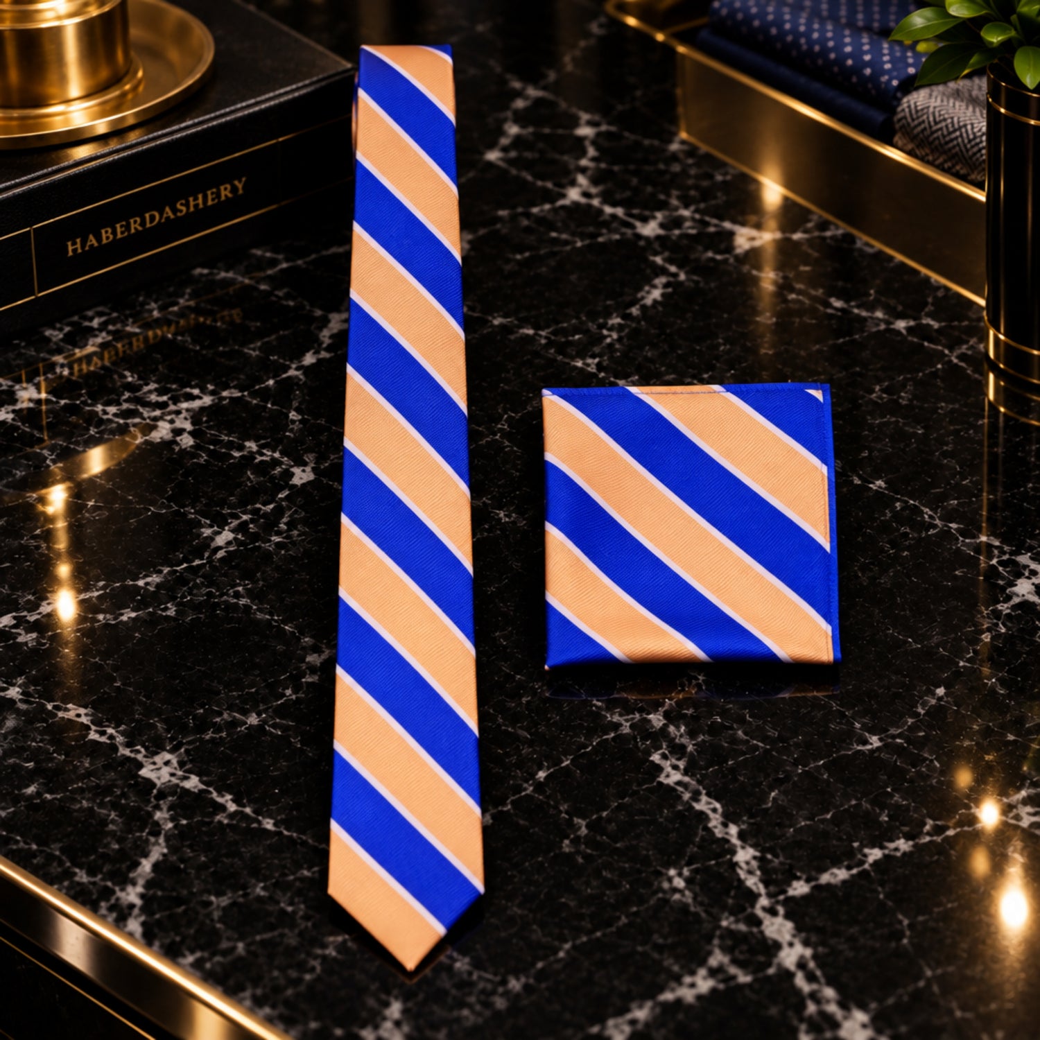 A men's necktie and matching pocket square set with diagonal stripes in royal blue, tan, and thin white lines, styled on a white dress shirt and black suit jacket.