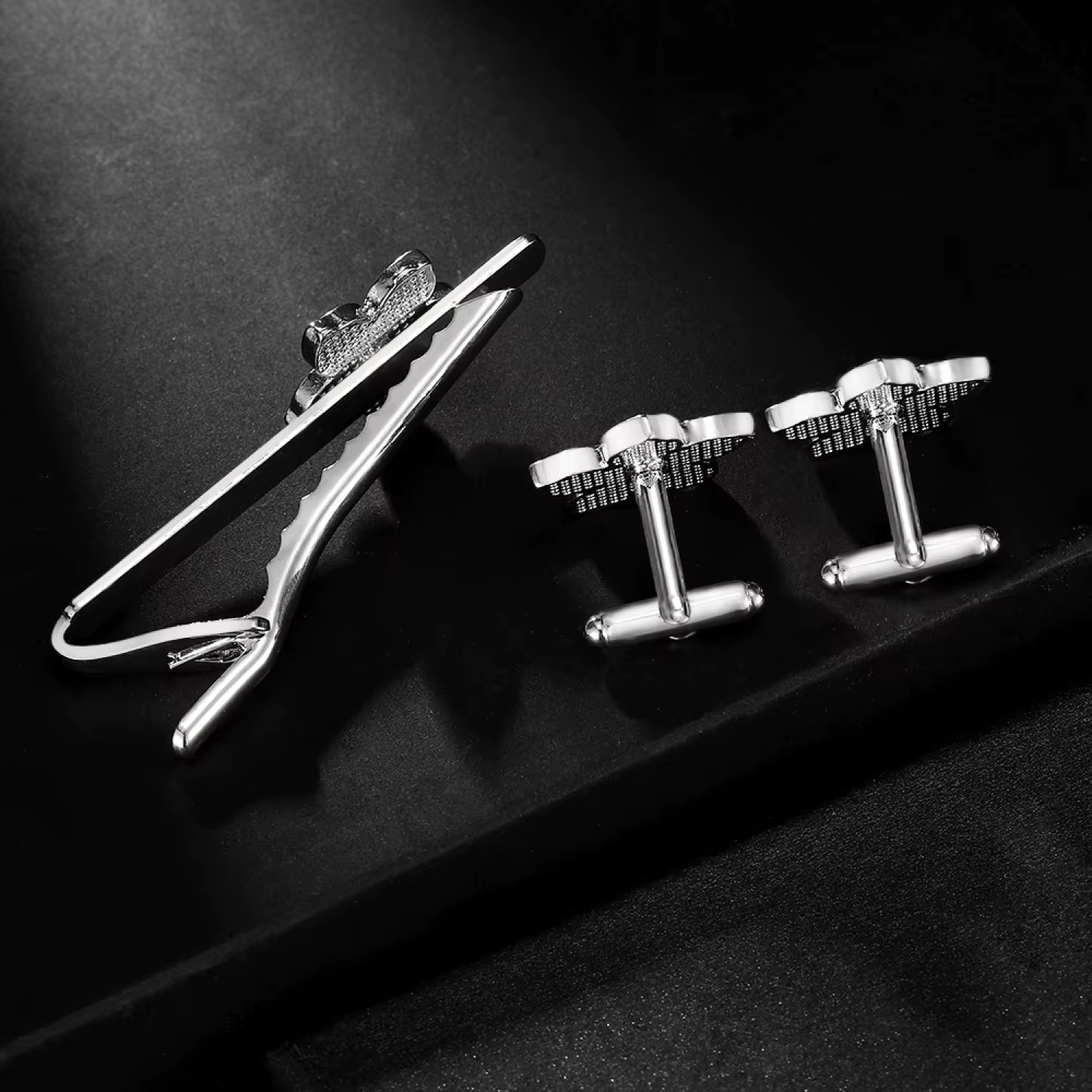 A silver-toned men's accessory set featuring a tie clip and matching cufflinks, displayed on a dark textured surface.
