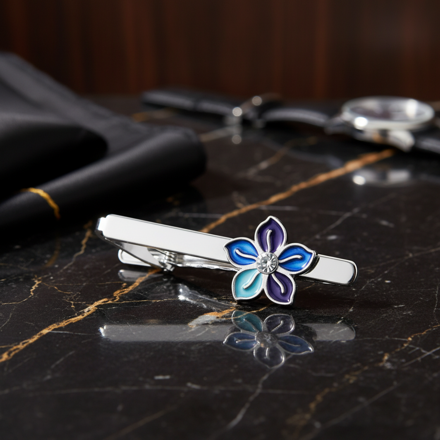 A silver-toned tie bar clip featuring a five-petal flower design with blue, purple, and light blue enamel petals and a small clear rhinestone in the center, shown on a black reflective background.
