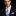 A man wearing a dark navy suit jacket, white dress shirt, a light blue and black striped tie, and a matching light blue pocket square, set against a solid black background.