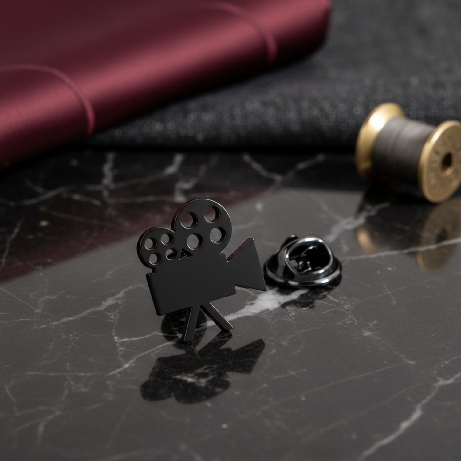 A black metal enamel pin shaped like a film camera, shown next to its silver metal button clasp on a dark textured fabric background.