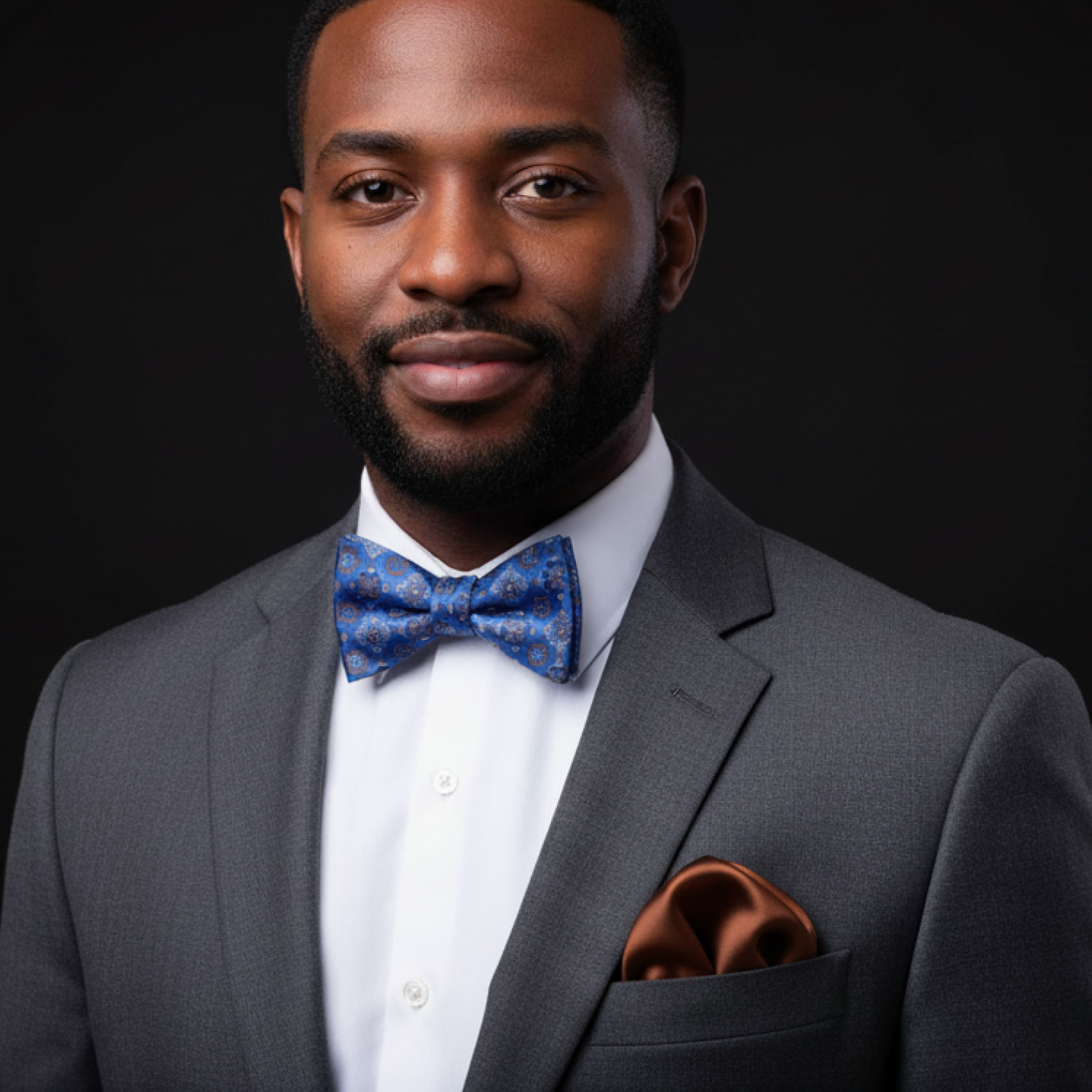 Man wearing a gray suit with a blue bow tie and brown pocket square on a black background