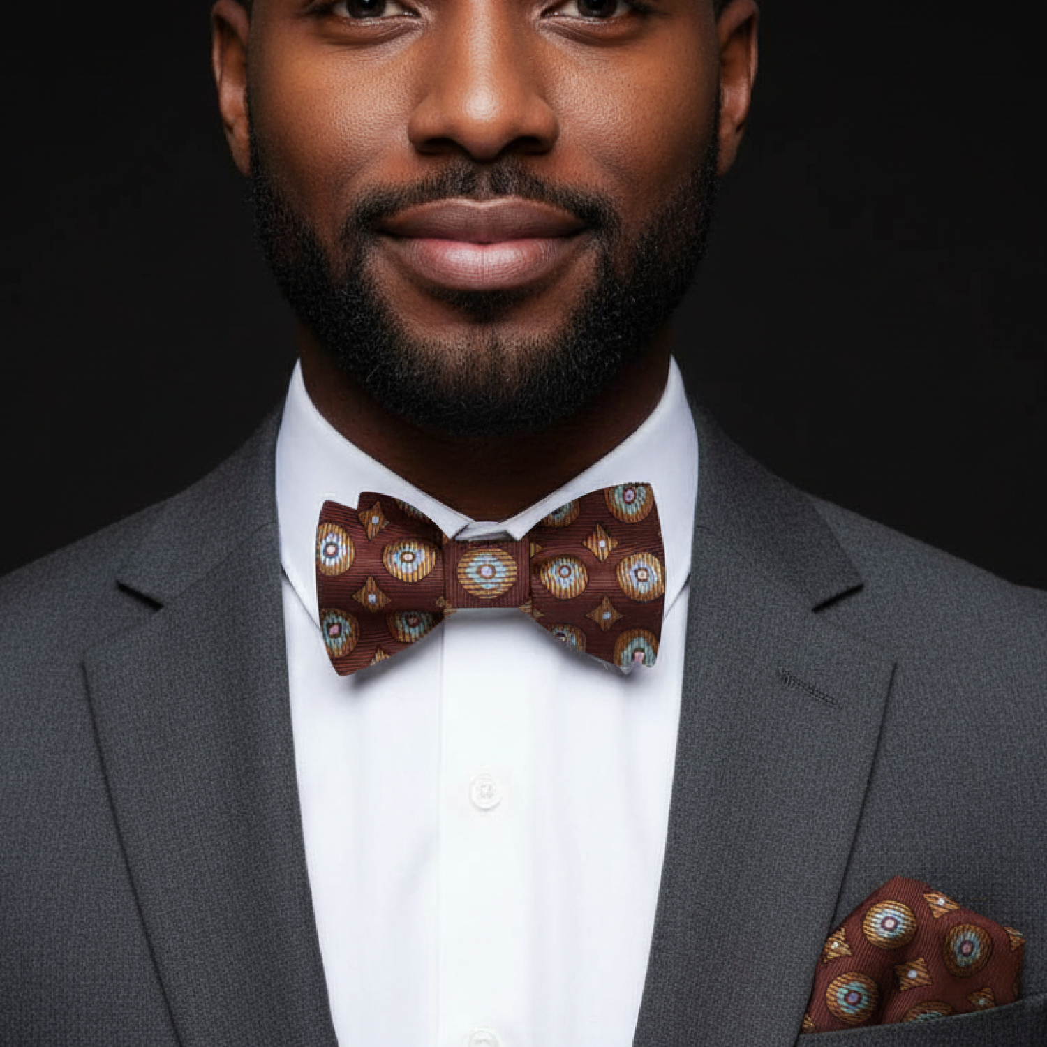 Man wearing a gray suit with a patterned bow tie and pocket square on a black background