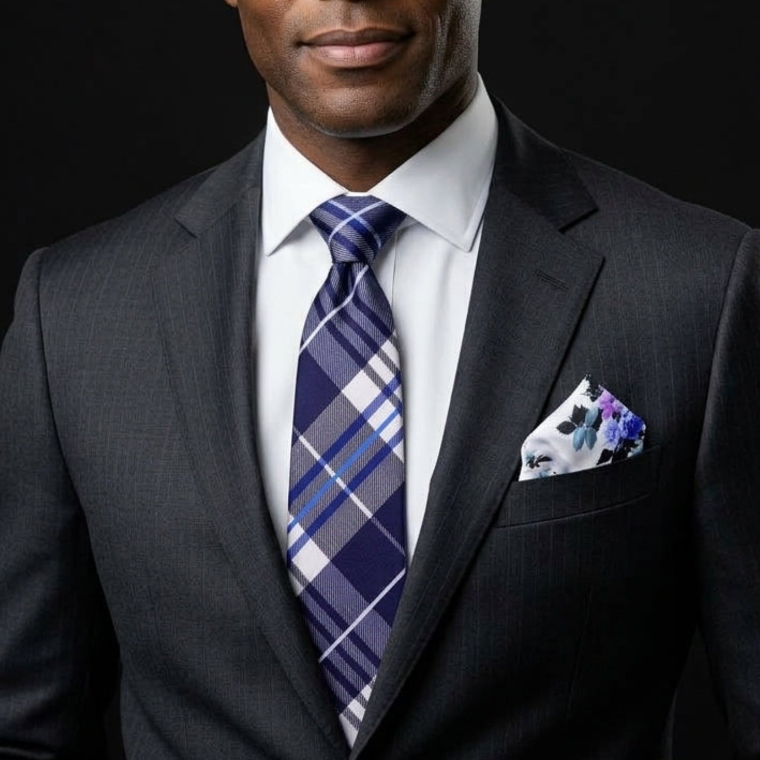 A man wearing a dark gray pinstripe suit, white dress shirt, a blue and purple plaid tie, and a matching pocket square, photographed against a solid black background.