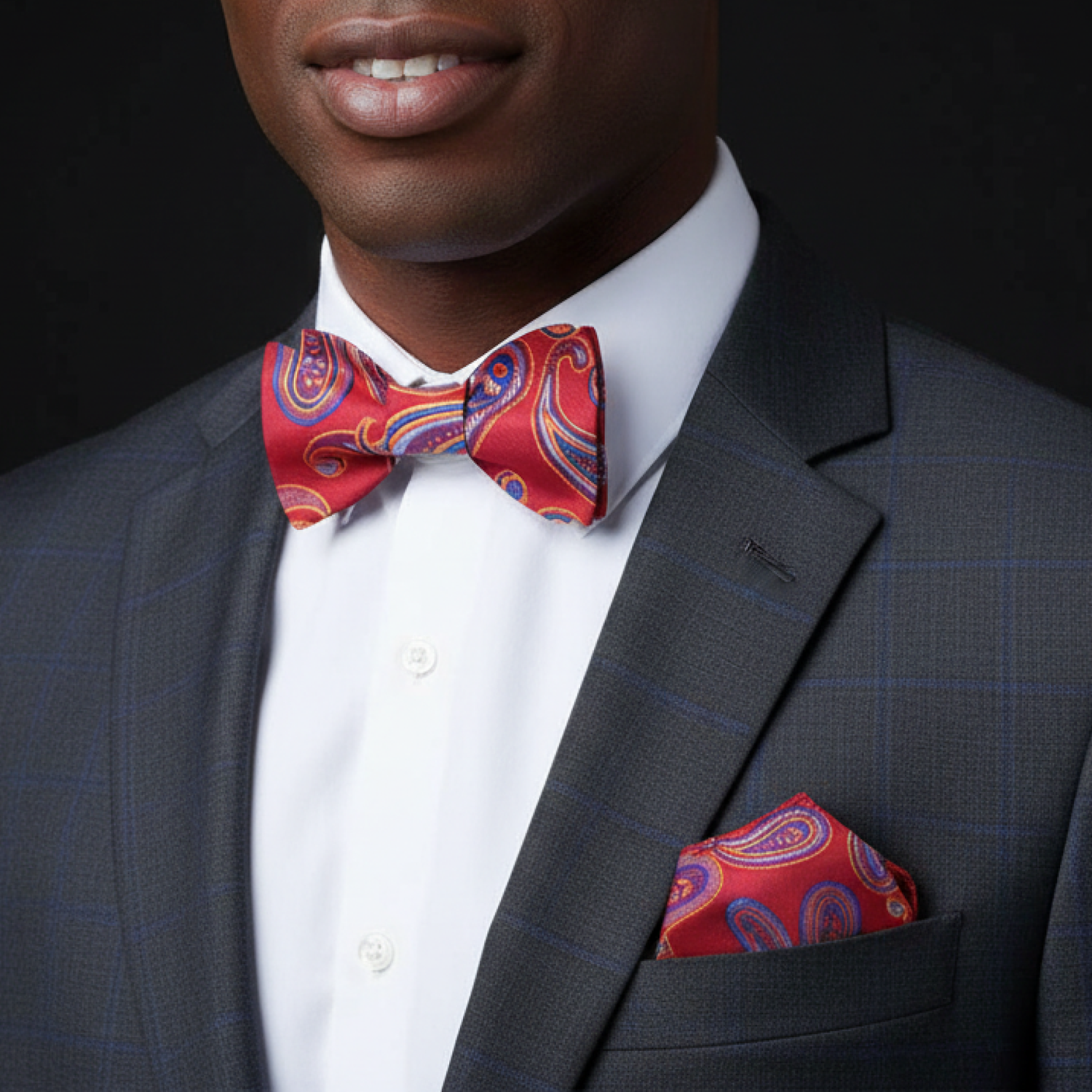 Man wearing a gray suit with a red paisley bow tie and pocket square against a black background