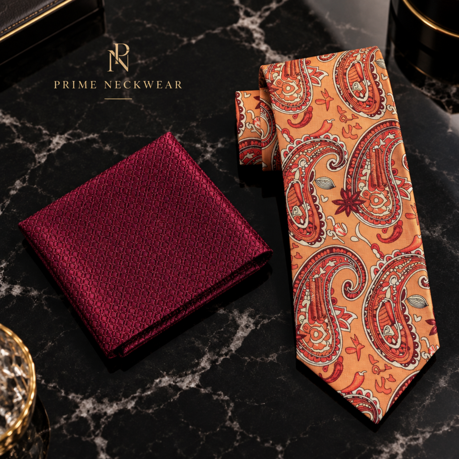 A men's necktie and matching pocket square set from Prime Neckwear, displayed on a black marble surface. The pocket square is a deep burgundy red with a subtle geometric pattern, while the necktie features a vibrant orange and red paisley pattern with black outlines.